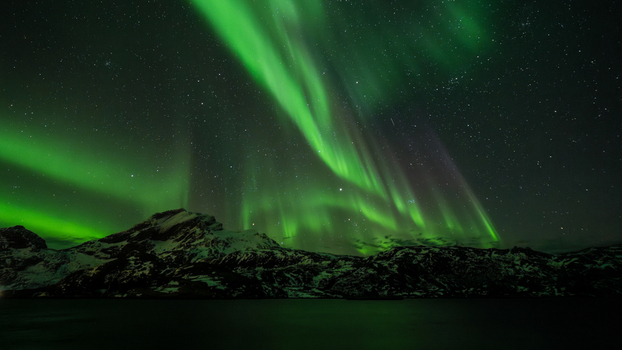 View of vibrant green aurora borealis dancing above the dark snow dusted mountain peaks and calm waters in Leknes Lofoten Norway.