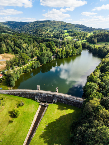 Aerial view of vodni nadrz fojtka reservoir with its stone dam surrounded by lush green forests and rolling hills under a blue sky in Mnisek Liberecky kraj Czechia. Aerial view of vodni nadrz fojtka reservoir with its stone dam surrounded by lush green forests and rolling hills under a blue sky in Mnisek Liberecky kraj Czechia.