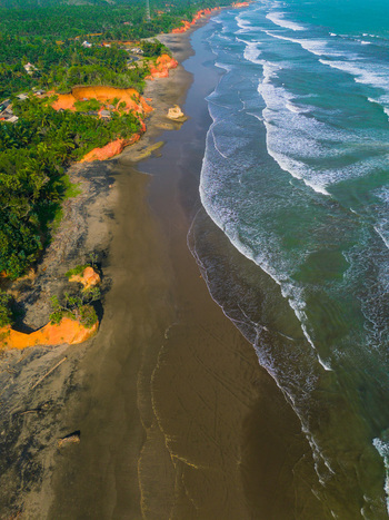 Aerial view of the rugged coastline with dark sand beach dramatic orange cliffs lush green vegetation and turquoise ocean waves in Bali Indonesia. Aerial view of the rugged coastline with dark sand beach dramatic orange cliffs lush green vegetation and turquoise ocean waves in Bali Indonesia.