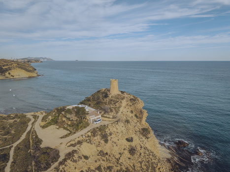 Aerial view of Torre del Xarco a historic stone watchtower on a rugged cliff overlooking the Mediterranean Sea with a small house and dirt paths in La Vila Joiosa Comunidad Valenciana Spain.