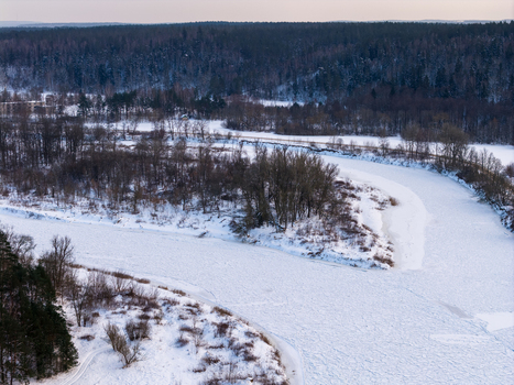 Aerial view of a frozen river winding through a snow covered forest landscape with bare trees under a soft winter sky Lithuania. Aerial view of a frozen river winding through a snow covered forest landscape with bare trees under a soft winter sky Lithuania.