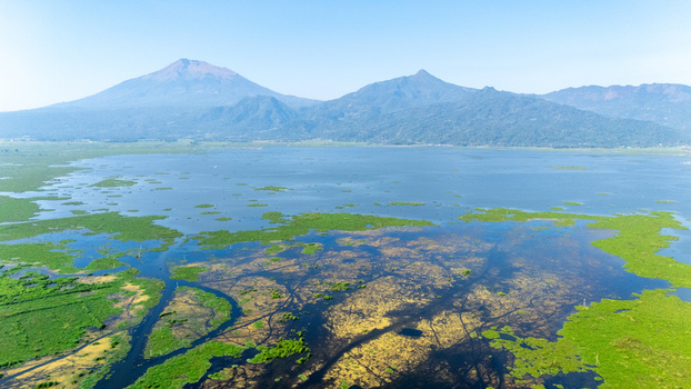 Aerial view of Lake Rawa Pening with lush green vegetation water channels and distant mountains under a clear blue sky in Waduk Rawapening Central Java Indonesia. Aerial view of Lake Rawa Pening with lush green vegetation water channels and distant mountains under a clear blue sky in Waduk Rawapening Central Java Indonesia.