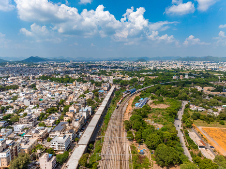 Aerial view of railway tracks cutting through the urban landscape surrounded by lush greenery and distant hills under a bright blue sky in Udaipur Rajasthan India. Aerial view of railway tracks cutting through the urban landscape surrounded by lush greenery and distant hills under a bright blue sky in Udaipur Rajasthan India.