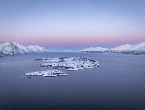 Aerial view of snow covered islands in a calm fjord surrounded by majestic mountains under a soft pink and blue twilight sky in Tromso Troms Norway. Aerial view of snow covered islands in a calm fjord surrounded by majestic mountains under a soft pink and blue twilight sky in Tromso Troms Norway.