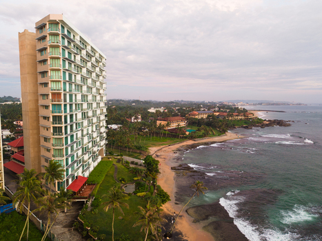Aerial view of a modern high rise building on the coastline of Gintota beach with palm trees and ocean waves crashing on the shore in Galle Southern Province Sri Lanka. Aerial view of a modern high rise building on the coastline of Gintota beach with palm trees and ocean waves crashing on the shore in Galle Southern Province Sri Lanka.