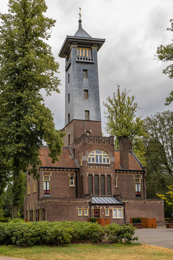 View of De Brandtoren a historic brick fire watchtower with a slate covered spire surrounded by lush green trees under a cloudy sky in Esbeek Noord Brabant Netherlands.