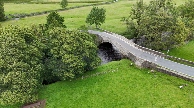 12 July 2025 Aerial view of a stone arch bridge spanning a dark river surrounded by vibrant green pastures stone walls and lush trees. 12 July 2025 Aerial view of a stone arch bridge spanning a dark river surrounded by vibrant green pastures stone walls and lush trees.