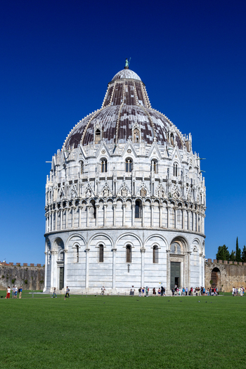 View of the Pisa Baptistery with its intricate marble facade and dome under a clear blue sky surrounded by a green lawn with people walking in Pisa Toscana Italy. View of the Pisa Baptistery with its intricate marble facade and dome under a clear blue sky surrounded by a green lawn with people walking in Pisa Toscana Italy.
