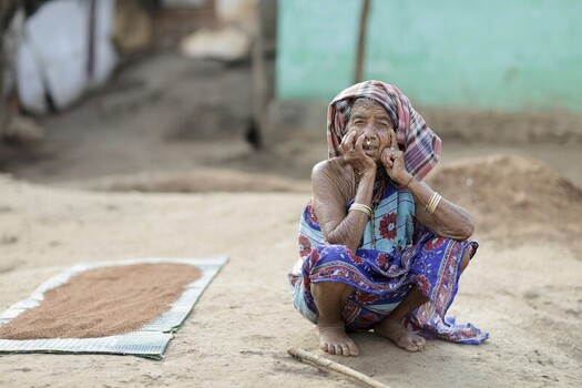 Odisha India 26 March 2026 View of an elderly woman with a patterned headscarf and traditional jewelry crouched on the sandy ground near a woven mat laden with grains. Odisha India 26 March 2026 View of an elderly woman with a patterned headscarf and traditional jewelry crouched on the sandy ground near a woven mat laden with grains.