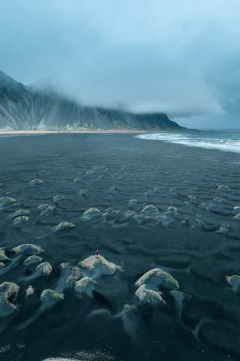 Aerial view of Vestrahorn mountain rising above the black sand beach with a person in a red coat standing on a dune under a cloudy sky in Hornafjordur Iceland. Aerial view of Vestrahorn mountain rising above the black sand beach with a person in a red coat standing on a dune under a cloudy sky in Hornafjordur Iceland.