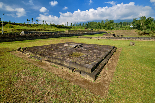 View of the ancient stone foundations of Situs Liyangan surrounded by green grass and stone walls under a blue sky in Temanggung Central Java Indonesia.