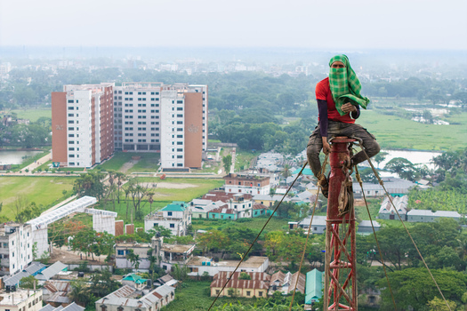 Trishal Upazila Bangladesh 13 May 2026 Aerial view of a worker with his face covered sitting on top of a high metal tower overlooking the landscape and modern buildings. Trishal Upazila Bangladesh 13 May 2026 Aerial view of a worker with his face covered sitting on top of a high metal tower overlooking the landscape and modern buildings.
