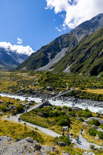 View of a winding path leading towards a bridge over a rushing river in a valley surrounded by lush green hills and snow capped mountains Mount Cook Canterbury Region New Zealand. View of a winding path leading towards a bridge over a rushing river in a valley surrounded by lush green hills and snow capped mountains Mount Cook Canterbury Region New Zealand.