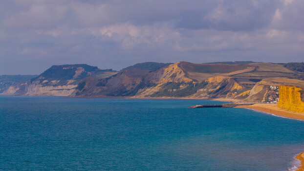 Aerial view of the dramatic golden sandstone cliffs of West Bay meeting the blue waters of the English Channel under a cloudy sky West Bay England United Kingdom. Aerial view of the dramatic golden sandstone cliffs of West Bay meeting the blue waters of the English Channel under a cloudy sky West Bay England United Kingdom.