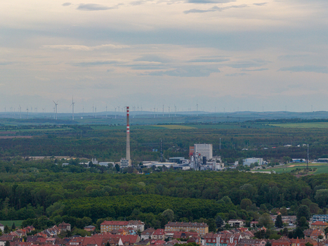 Aerial view of an industrial complex with a tall chimney and wind turbines on the horizon beyond a lush green forest in Breclav South Moravian Region Czechia.