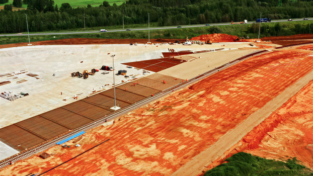Aerial view of a large construction site featuring red soil concrete foundations and rebar grids with workers present near a highway in Latvia. Aerial view of a large construction site featuring red soil concrete foundations and rebar grids with workers present near a highway in Latvia.