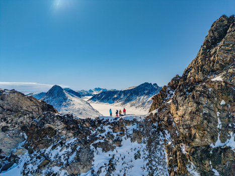 Aerial view of a group of people standing on a snowy mountain ridge overlooking a vast glacier valley and rugged peaks under a clear blue sky in Tasiilaq Sermersooq Municipality Greenland. Aerial view of a group of people standing on a snowy mountain ridge overlooking a vast glacier valley and rugged peaks under a clear blue sky in Tasiilaq Sermersooq Municipality Greenland.