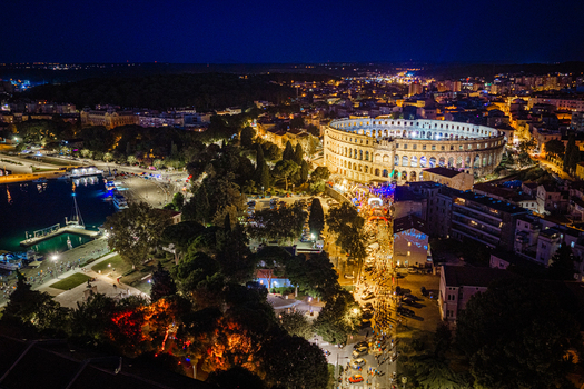 Aerial view of the illuminated Pula Arena at night surrounded by the historic city streets harbor lights and lush trees in Pula Istria County Croatia. Aerial view of the illuminated Pula Arena at night surrounded by the historic city streets harbor lights and lush trees in Pula Istria County Croatia.