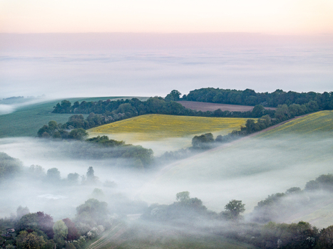 Aerial view of rolling green hills and lush fields partially covered by thick morning mist under a soft dawn sky Nuffield Oxfordshire United Kingdom. Aerial view of rolling green hills and lush fields partially covered by thick morning mist under a soft dawn sky Nuffield Oxfordshire United Kingdom.