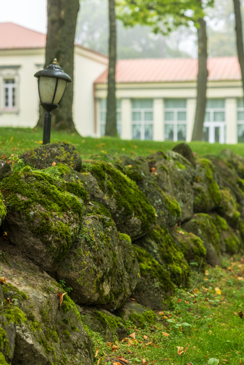 View of a mossy stone wall and a vintage lamp post in front of the historic Taujenai Manor surrounded by lush green trees in Taujenai Vilnius County Lithuania.