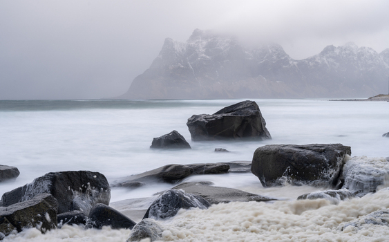 View of the rocky coastline at Leknes with sea foam on the sand large boulders in the foreground and misty snow capped mountains in Leknes Lofoten Norway.