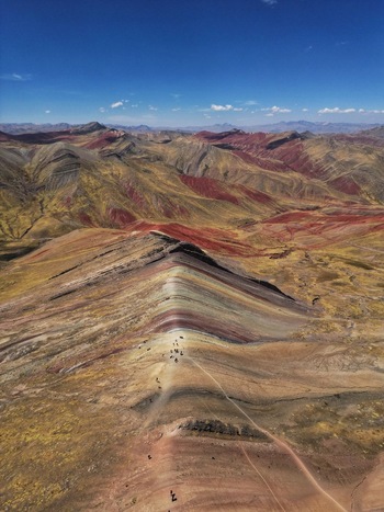 Aerial view of Vinicunca Rainbow Mountain showing vibrant mineral stripes and hikers on the ridge under a clear blue sky in Quispicanchi Province Cusco Peru.