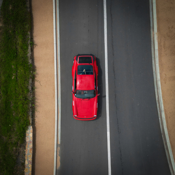 Aerial view of a red vintage sports car driving on a paved road with white markings and sandy shoulders in La Chapelle de Guinchay Bourgogne Franche Comte France.