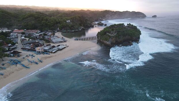 Aerial view of Pantai Drini featuring a sandy beach rocky islet fishing boats and turquoise waves crashing against the coast in Kecamatan Tanjungsari Daerah Istimewa Yogya Indonesia. Aerial view of Pantai Drini featuring a sandy beach rocky islet fishing boats and turquoise waves crashing against the coast in Kecamatan Tanjungsari Daerah Istimewa Yogya Indonesia.