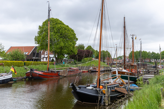 View of traditional wooden sailing boats moored at Hafen Harlesiel with lush green trees and houses under a cloudy sky in Wittmund Lower Saxony Germany. View of traditional wooden sailing boats moored at Hafen Harlesiel with lush green trees and houses under a cloudy sky in Wittmund Lower Saxony Germany.