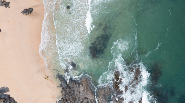 Aerial view of Harlyn Bay Beach with turquoise ocean waves crashing against dark jagged rocks and golden sand in Saint Merryn Cornwall United Kingdom.