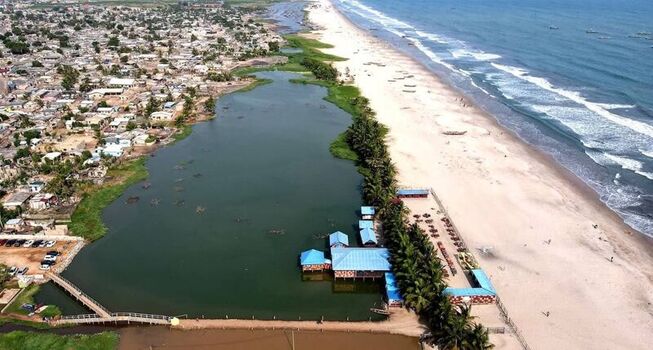 Aerial view of a coastal lagoon adjacent to a sandy beach and the Atlantic Ocean with a nearby town featuring residential buildings and palm trees in Ghana. Aerial view of a coastal lagoon adjacent to a sandy beach and the Atlantic Ocean with a nearby town featuring residential buildings and palm trees in Ghana.