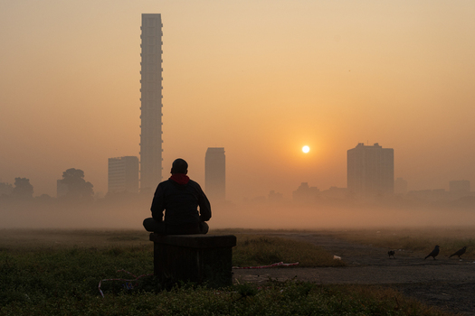 View of a person sitting on a bench in a park overlooking the hazy city skyline during sunrise in Kolkata West Bengal India. View of a person sitting on a bench in a park overlooking the hazy city skyline during sunrise in Kolkata West Bengal India.