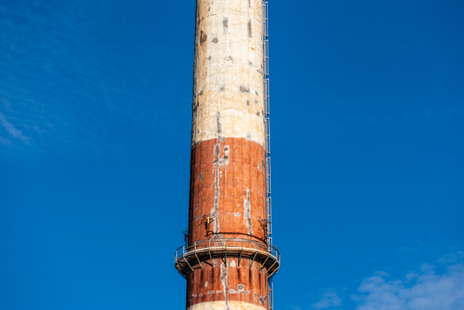 View of a tall industrial chimney with red and white painted concrete a vertical ladder and a metal maintenance platform against a clear blue sky in Poland.