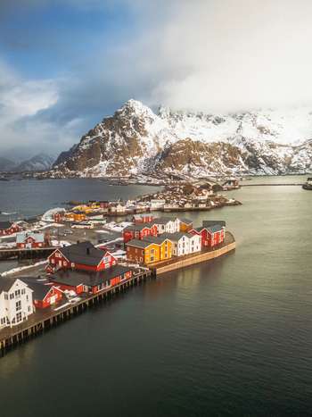 Aerial view of vibrant red and yellow cabins contrast against the snow dusted peaks and tranquil waters of the Lofoten Islands Henningsv&aelig;r Nordland Norway.