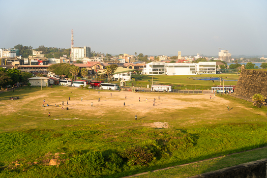 View of people playing cricket on a grassy field near the historic fort ramparts and urban buildings Galle Southern Province Sri Lanka.