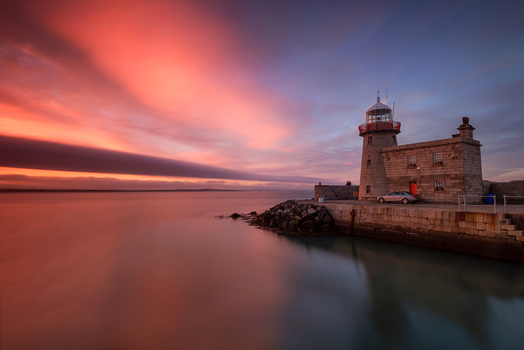 View of the historic Howth Harbour lighthouse at sunset with vibrant orange and pink clouds over calm water Ireland. View of the historic Howth Harbour lighthouse at sunset with vibrant orange and pink clouds over calm water Ireland.