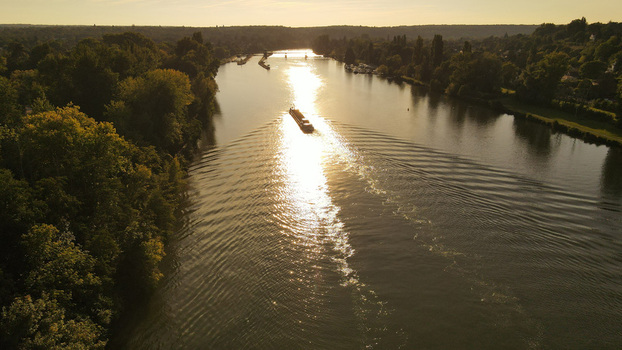 Aerial view of a barge navigating the Seine river during golden hour surrounded by lush green trees along the banks in Chartrettes Ile de France France. Aerial view of a barge navigating the Seine river during golden hour surrounded by lush green trees along the banks in Chartrettes Ile de France France.