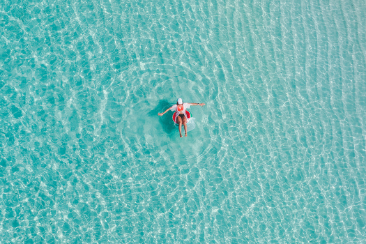 Aerial view of a person wearing a white hat and shirt floating on a red inflatable ring in crystal clear turquoise water with sandy ripples in Vashafaru Haa Alif Atoll Maldives. Aerial view of a person wearing a white hat and shirt floating on a red inflatable ring in crystal clear turquoise water with sandy ripples in Vashafaru Haa Alif Atoll Maldives.