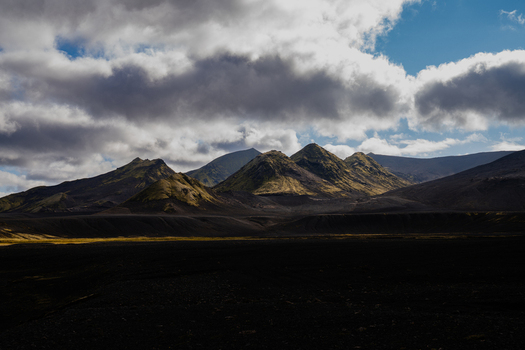 View of jagged peaks some touched by sunlight rise from dark volcanic earth under a sky streaked with dramatic clouds Skaftárhreppur Iceland. View of jagged peaks some touched by sunlight rise from dark volcanic earth under a sky streaked with dramatic clouds Skaftárhreppur Iceland.