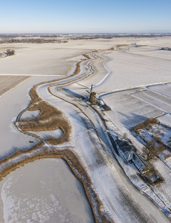 Aerial view of a traditional Dutch windmill standing beside a winding frozen canal in a snow covered landscape under a clear winter sky in Ursem North Holland Netherlands.