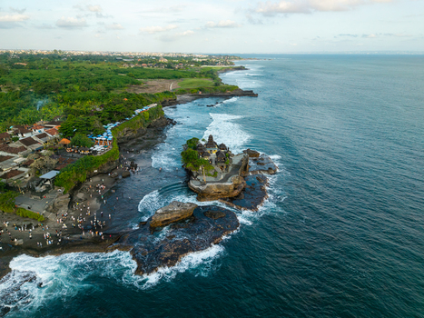 Aerial view of Tanah Lot Temple on a rocky outcrop surrounded by the Indian Ocean with crashing waves lush greenery and tourists on the beach in Kecamatan Kediri Bali Indonesia. Aerial view of Tanah Lot Temple on a rocky outcrop surrounded by the Indian Ocean with crashing waves lush greenery and tourists on the beach in Kecamatan Kediri Bali Indonesia.