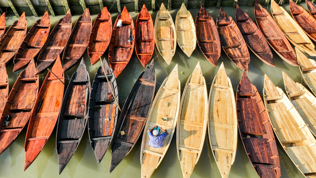 Aerial view of a fleet of wooden boats arrayed in a spectrum of earthy hues resting on the still olive green waters of Pirojpur Pourashava Pirojpur Pourashava Barisal Division Bangladesh. Aerial view of a fleet of wooden boats arrayed in a spectrum of earthy hues resting on the still olive green waters of Pirojpur Pourashava Pirojpur Pourashava Barisal Division Bangladesh.