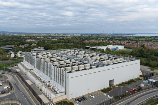 Aerial view of a large modern data center facility featuring numerous cooling units on the roof surrounded by parking areas access roads and suburban landscape United Kingdom.