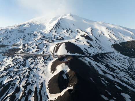 Aerial view of the southern slope of Mount Etna with snow covered volcanic craters and winding mountain roads under a clear blue sky in Nicolosi Sicilia Italy. Aerial view of the southern slope of Mount Etna with snow covered volcanic craters and winding mountain roads under a clear blue sky in Nicolosi Sicilia Italy.