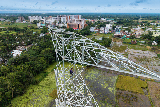 Trishal Upazila Bangladesh 13 May 2026 Aerial view of a high voltage electricity transmission tower under construction with a worker climbing the metal structure above green fields. Trishal Upazila Bangladesh 13 May 2026 Aerial view of a high voltage electricity transmission tower under construction with a worker climbing the metal structure above green fields.