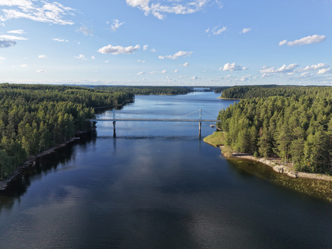 Aerial view of Potkusalmen silta Bridge spanning the calm waters of a lake surrounded by dense pine forests under a clear blue sky in Savonlinna Etela Savo Finland.