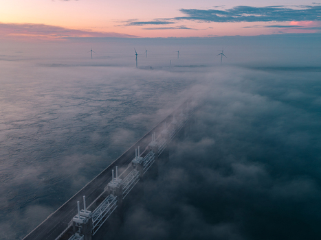 Aerial view of the Oosterscheldekering storm surge barrier shrouded in thick fog with wind turbines under a colorful dawn sky Vrouwenpolder Zeeland Netherlands.