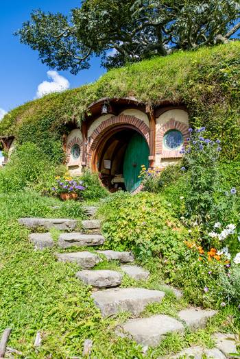 View of a quaint Hobbit hole nestled into a verdant hillside with a round green door and stone steps in Hobbiton Movie Set Matamata New Zealand. View of a quaint Hobbit hole nestled into a verdant hillside with a round green door and stone steps in Hobbiton Movie Set Matamata New Zealand.