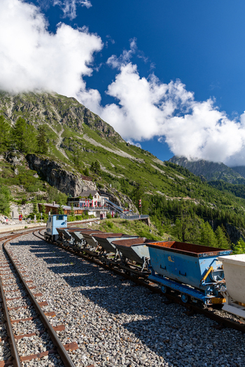 View of VerticAlp Emosson mountain railway with vintage mining carts on tracks set against a backdrop of lush green alpine slopes and a clear blue sky in Le Chatelard Fribourg Switzerland. View of VerticAlp Emosson mountain railway with vintage mining carts on tracks set against a backdrop of lush green alpine slopes and a clear blue sky in Le Chatelard Fribourg Switzerland.