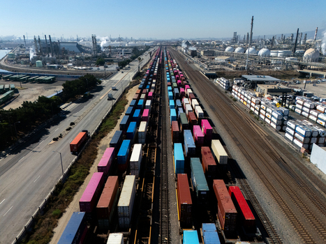 Aerial view of a long freight train loaded with colorful shipping containers traveling through an industrial area with storage tanks and refineries in Carson California United States.